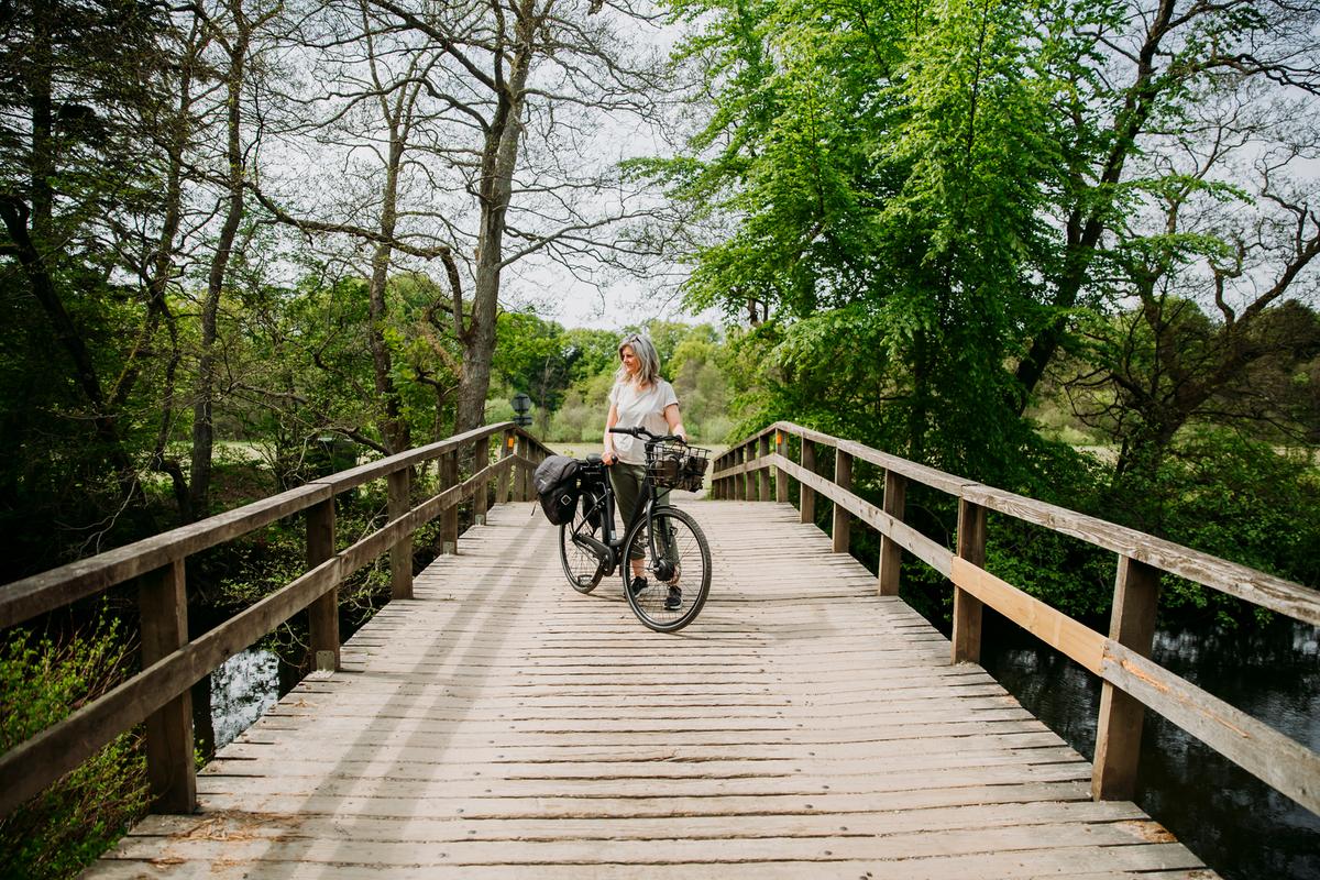 Kvinde står med en sort elcykel på en træbro ved Gudenåen nær Den Genfundne Bro